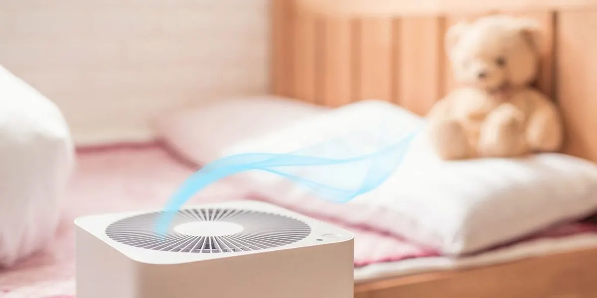 Weißer Luftreiniger mit blauem Luftstrom im Vordergrund, auf einem rosa Bett mit weißem Kissen und Teddybär im Hintergrund.