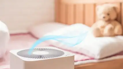 Weißer Luftreiniger mit blauem Luftstrom im Vordergrund, auf einem rosa Bett mit weißem Kissen und Teddybär im Hintergrund.