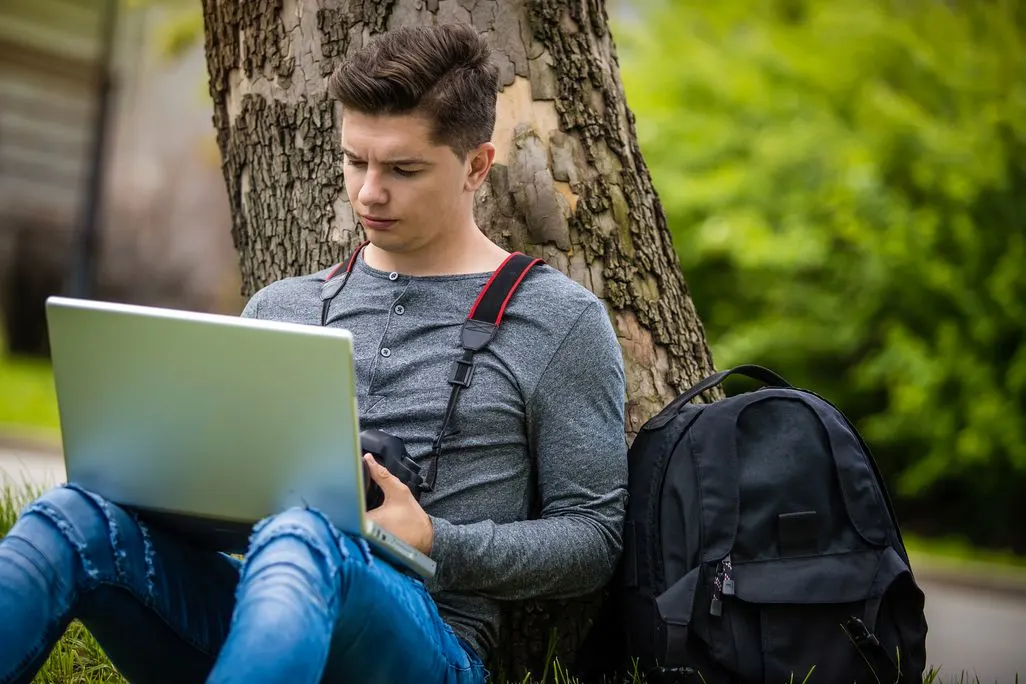 Mann arbeitet mit Laptop im Park unter einem Baum mit Rucksack neben sich