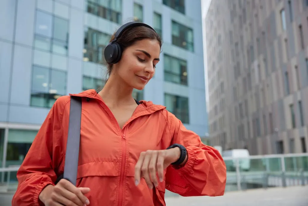 Frau mit orangener Jacke und schwarzen Kopfhörern sieht auf ihre Smartwatch.