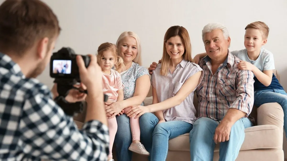 Ein Fotograf in kariertem Hemd macht ein Bild von einer lächelnden Familie (zwei Kinder, drei Erwachsene) auf einem hellen Sofa.