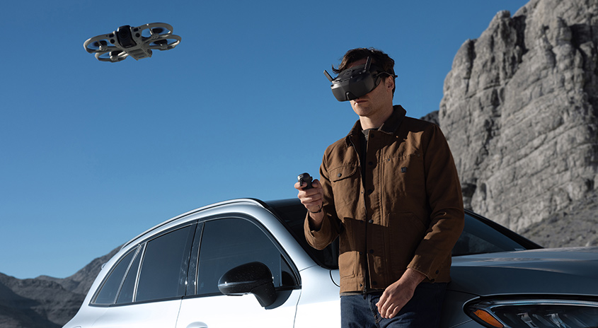 Person mit FPV-Brille steuert DJI Drohne neben geparktem Auto in felsiger Berglandschaft, klarer blauer Himmel, Fokus auf immersives Flugerlebnis.