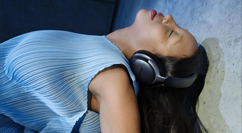 Eine Person liegt mit Kopfhörern auf dem Kopf. Sie trägt ein blaues, plissiertes Oberteil. Der Hintergrund ist dunkel.