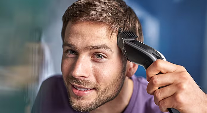 Ein Mann schneidet sich mit einem schwarzen Haarschneider die Haare vor einem blauen Hintergrund.