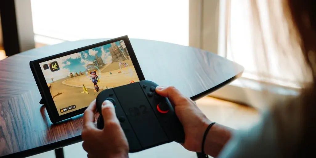 Eine Person spielt ein Videospiel auf einem Tablet mit einem schwarzen Controller mit roten Akzenten. Der Bildschirm zeigt ein Wüstenrennen mit einem Motorradfahrer und Bananen. Das Tablet steht auf einem dunklen Holztisch vor einem hellen Fenster.