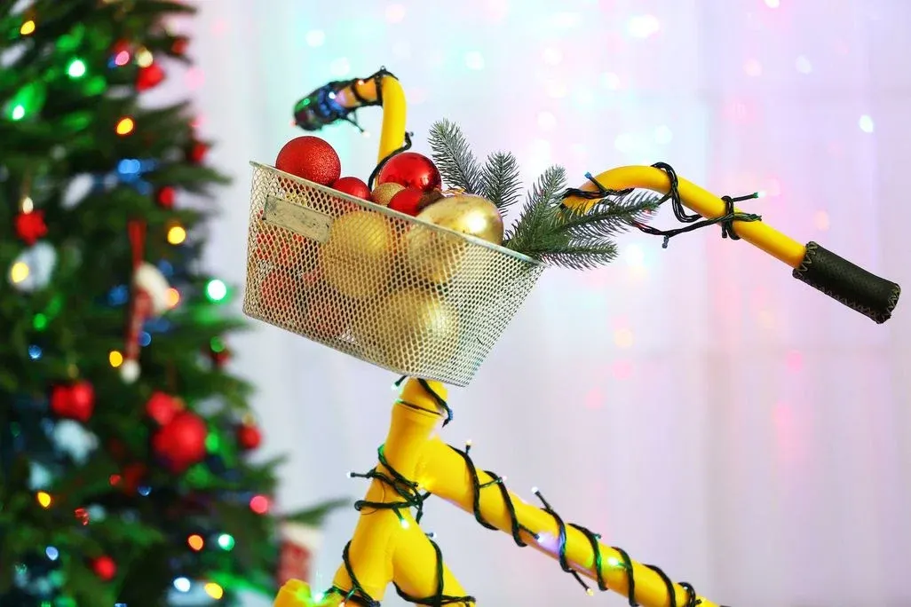 Gelber Fahrradrahmen mit Korb voller roter, goldener Kugeln und Tannenzweigen, geschmückt mit Lichterketten. Unscharfer Weihnachtsbaum im Hintergrund.