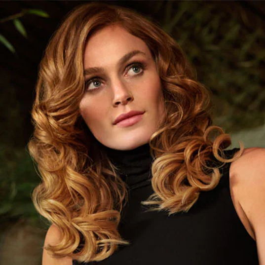 Eine Person mit gelocktem Haar trägt einen schwarzen Rollkragenpullover vor einem dunklen, unscharfen Hintergrund mit grünen Blättern.