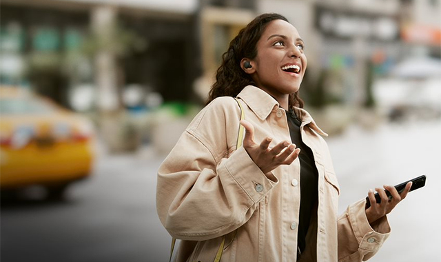 Eine Frau mit Kopfhörern hält ein schwarzes Smartphone in der Hand. Im Hintergrund ist eine gelbes Taxi und Gebäude zu sehen.