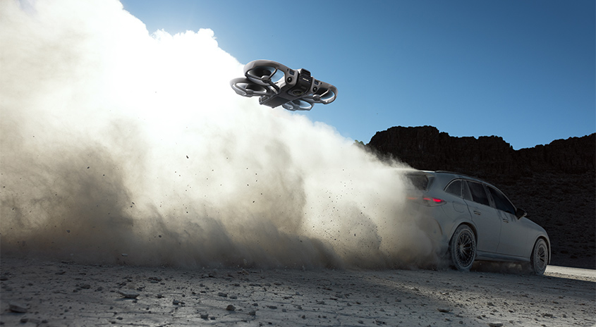  Drohne fliegt über staubige Wüstenpiste neben beschleunigendem Auto, riesige Staubwolke im Gegenlicht – dynamische Actionaufnahme in rauer Umgebung.