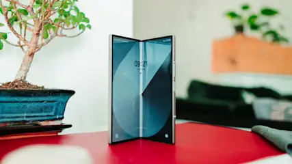 Ein aufgestelltes faltbares Smartphone steht auf einem roten Tisch neben einer Bonsai-Pflanze.