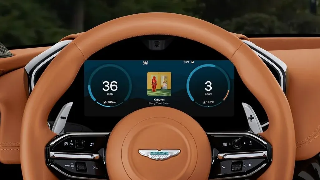 Braunes Lederlenkrad eines Aston Martin. Das digitale Display zeigt 36 mph, Fahrmodus Sport und ein Musikalbum-Cover.