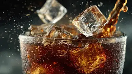 Glas mit sprudelndem Cola‑Getränk und Eiswürfeln, Flüssigkeit wird eingegossen und Tropfen spritzen.