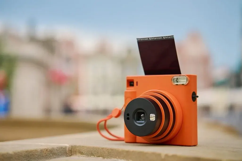 Orange Sofortbildkamera mit schwarzem Objektiv und aufgeklapptem dunkelrotem Blitz auf Sand vor unscharfem Strandszene-Hintergrund – Vintage-Fotografie und Urlaubserlebnis symbolisierend.