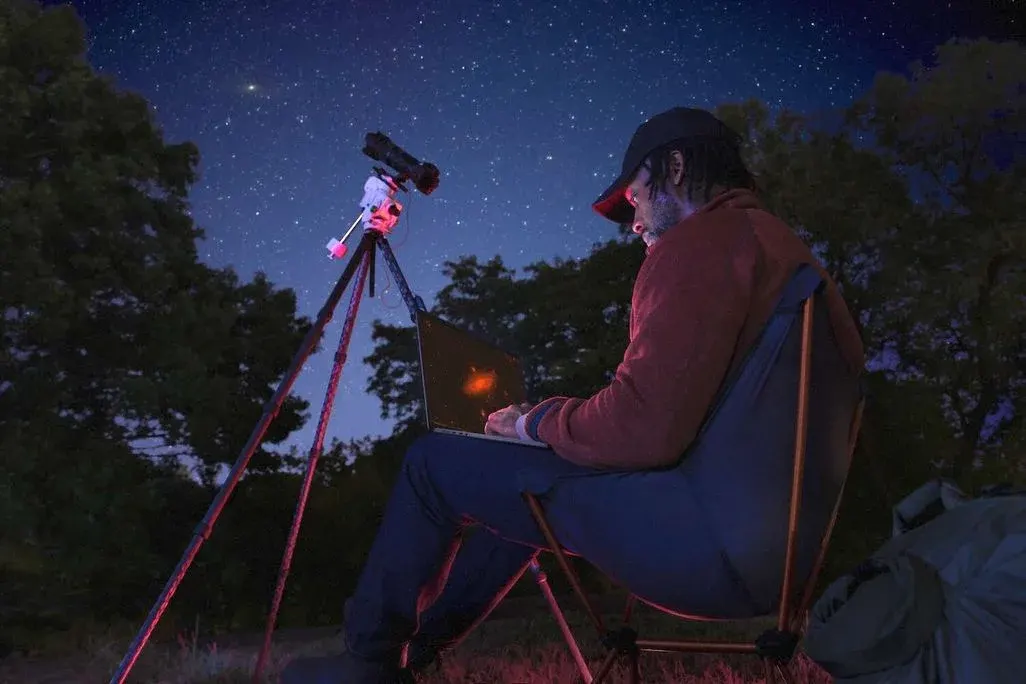Mann sitzt nachts mit einer Kamera samt Stativ und einem neuen MacBook-Pro am Schoß in der Natur, um den Himmel zu beobachten.