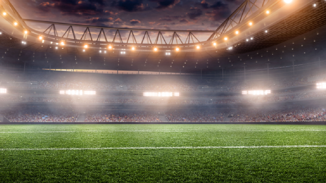 Ein beleuchtetes Fußballstadion mit grüner Grasfläche und weißen Linien. Im Hintergrund Tribünen und ein bewölkter Abendhimmel.