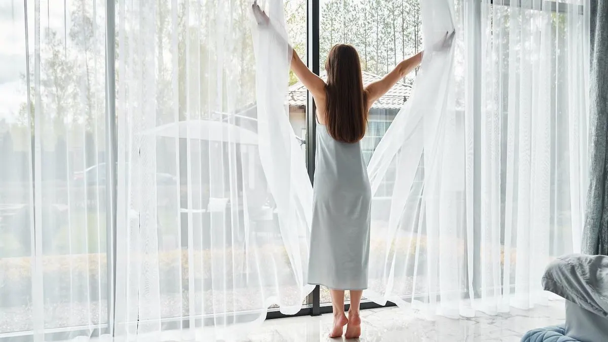 Frau öffnet morgens weiße Vorhänge vor einer großen Fensterfront.