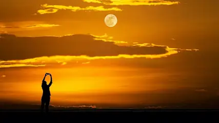 Person macht ein Foto vom Vollmond am orangefarbenen Himmel bei Sonnenuntergang.