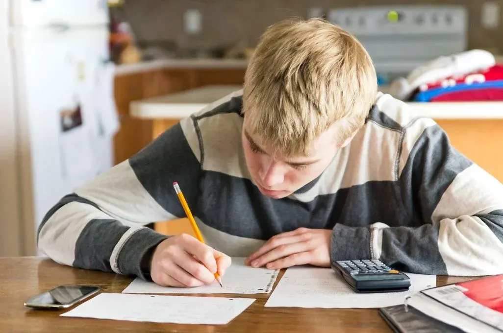 Jugendlicher sitzt am Tisch und löst Aufgaben; schreibt mit Bleistift auf Papier, neben ihm Taschenrechner und Handy.