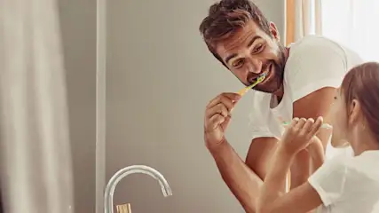 Erwachsener und Kind stehen gemeinsam vor einem Waschbecken im Badezimmer und putzen sich die Zähne.