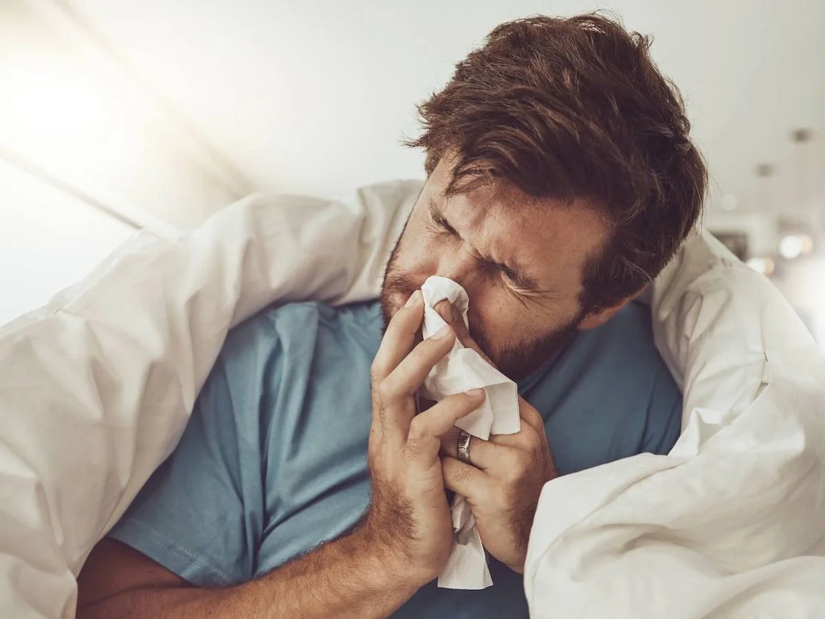 Krank wirkender Mann sitzt im Bett, in eine Decke gehüllt, und putzt sich mit einem Taschentuch die Nase.