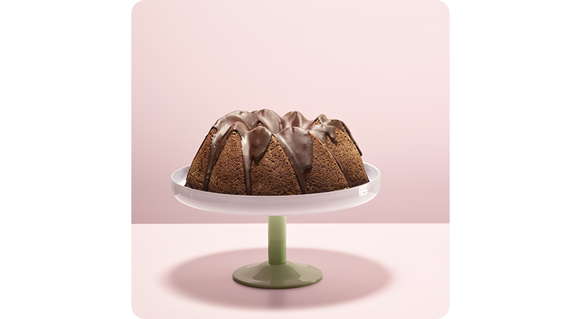 Gugelhupf mit Schokoglasur auf eleganter Tortenplatte mit grünem Fuß, vor pastellrosa Hintergrund stilvoll in Szene gesetzt.