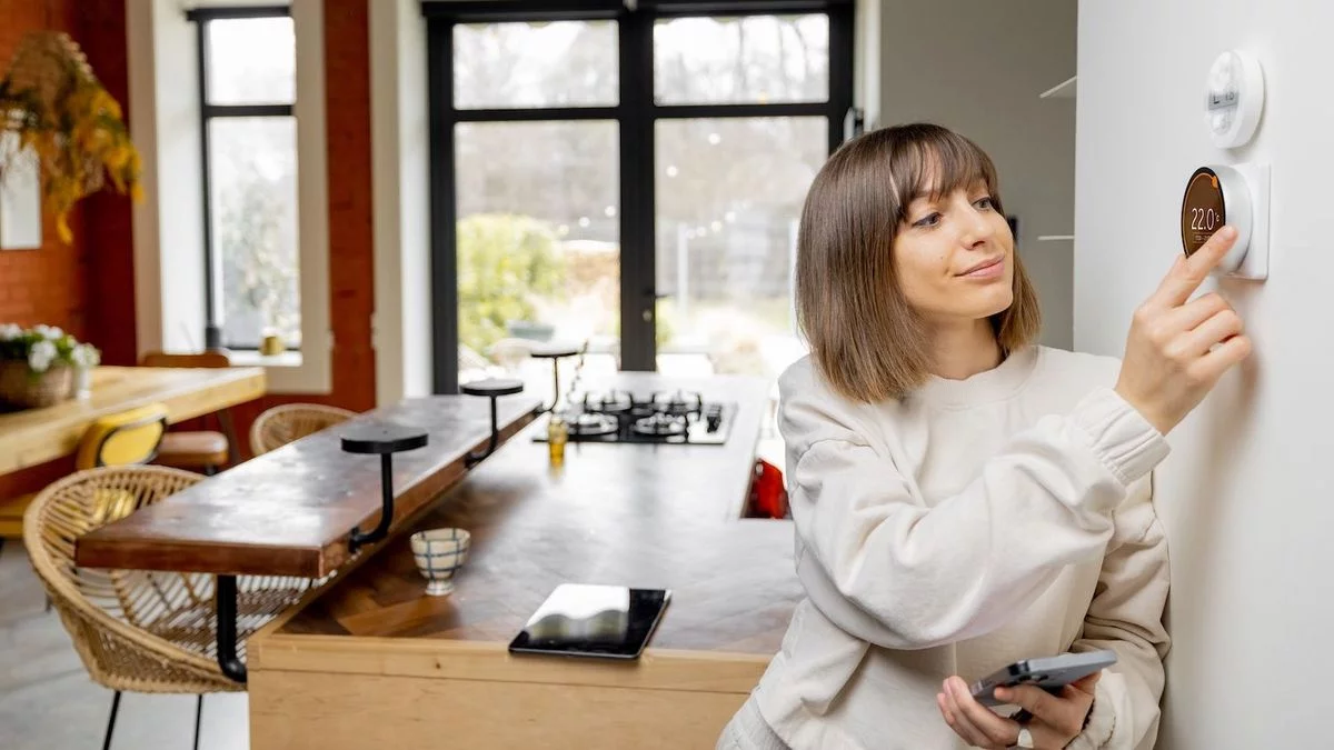 Frau in moderner Wohnküche stellt die Temperatur am smarten Wandthermostat ein und hält dabei ihr Smartphone in der Hand.