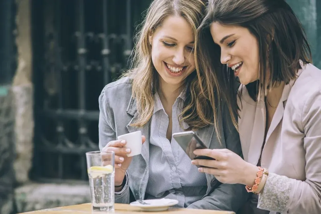 Zwei lächelnde Frauen an einem Holztisch, eine mit weißer Kaffeetasse, die andere mit Smartphone, blicken gemeinsam darauf. Ein Glas Wasser mit Zitrone steht daneben.