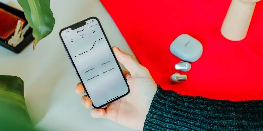 Ein Smartphone mit Equalizer-App, daneben liegen In-Ear-Kopfhörer und Ladecase.