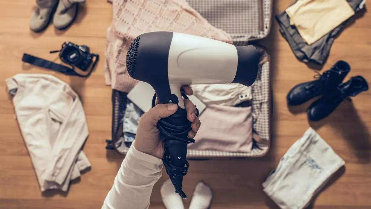 Eine Hand hält einen schwarz-weißen Haartrockner. Daneben ein offener Koffer mit Kleidung, Schuhen und Kamera auf Holzboden.