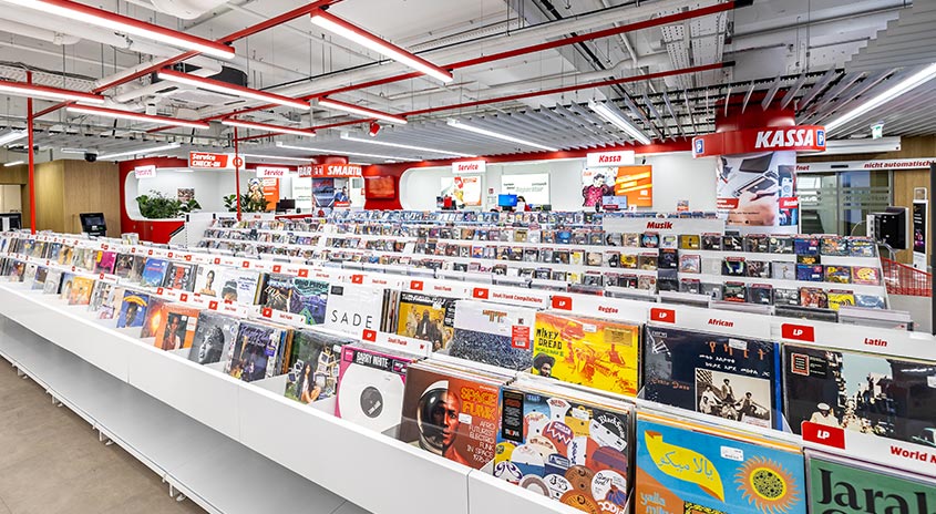 Ein Geschäft mit langen, weißen Regalen voller Schallplatten. Rote Akzente sind an der Decke und an Schildern zu sehen.