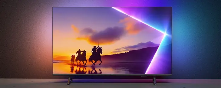 Flacher Fernseher mit eingeschaltetem Bildschirm vor dunkler Wand, darauf eine Szene von Reitern am Strand bei Sonnenuntergang; der TV ist von farbigem Ambient-Light in Lila und Blau hinterleuchtet.