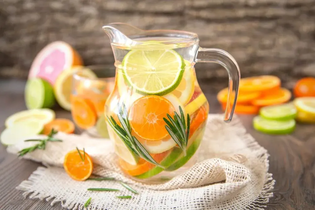 Klare Glaskaraffe mit Zitruswasser (Orange, Limette, Zitrone) und Rosmarin auf hellem Stoff. Geschnittene Früchte auf dunklem Holztisch.