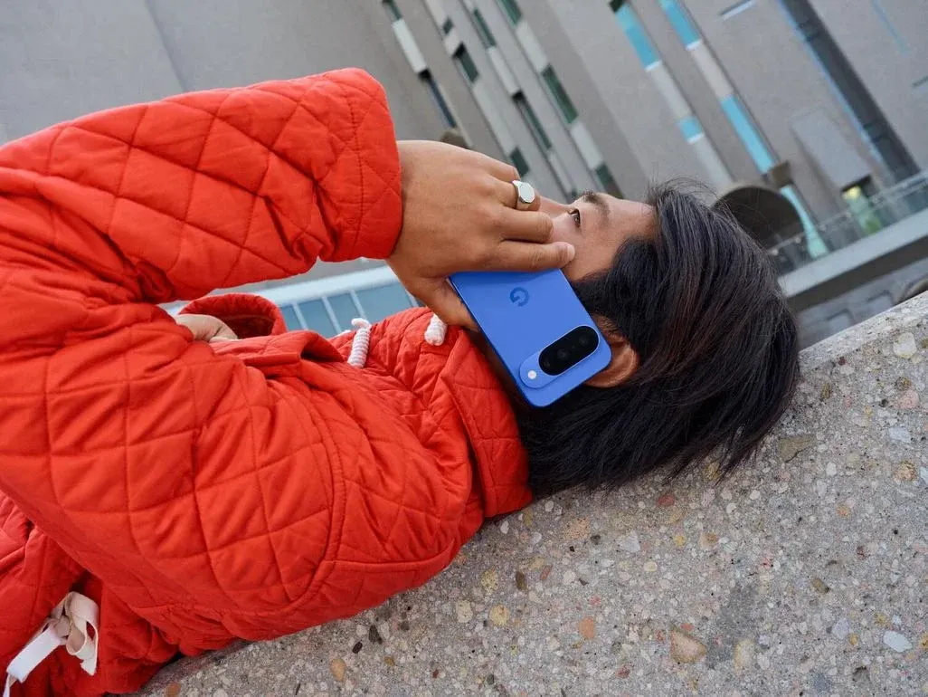 Person in roter Steppjacke telefoniert mit blauem Handy, trägt Ring, liegt auf Beton. Gebäude mit Fenstern im Hintergrund.