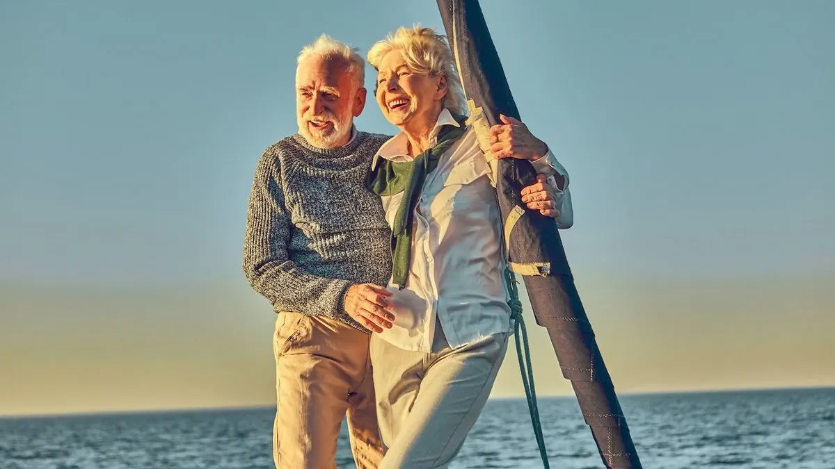 Zwei lachende ältere Erwachsene auf einem Boot. Der Mann trägt einen grauen Pullover, die Frau eine weiße Bluse und hält einen Mast. Blauer Himmel und Meer.