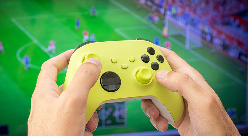 Eine Person hält einen gelben Controller vor einem Bildschirm, der ein Fußballspiel zeigt.