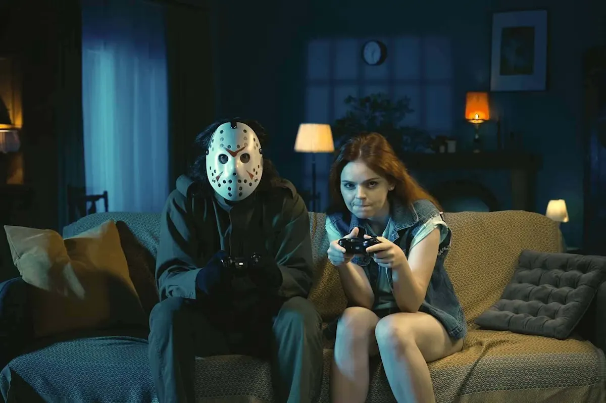 Ein Mann mit weißer Maske sitzt neben einem Mädchen in einem dunklen Wohnzimmer auf dem Sofa. Beide spielen mit Controllern ein Konsolengame.