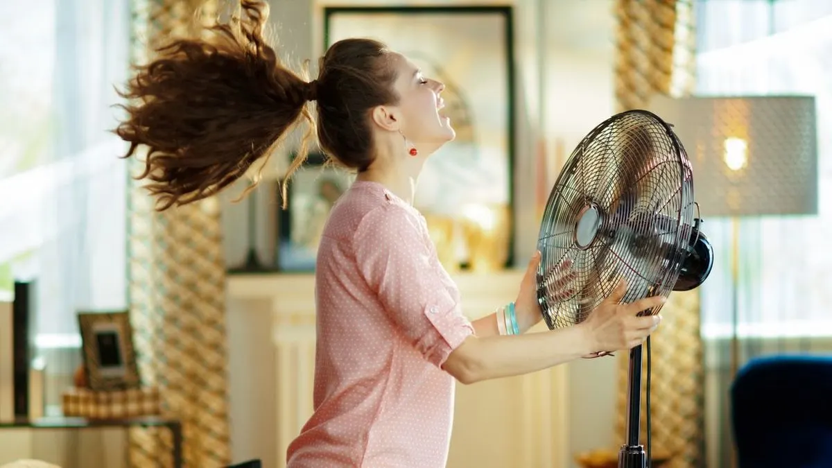 Frau mit langen Haaren steht vor einem laufenden Standventilator im Wohnzimmer