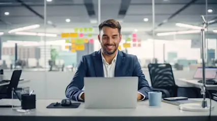 Mann im Anzug sitzt an einem Schreibtisch in einem modernen Buero und arbeitet am Laptop.