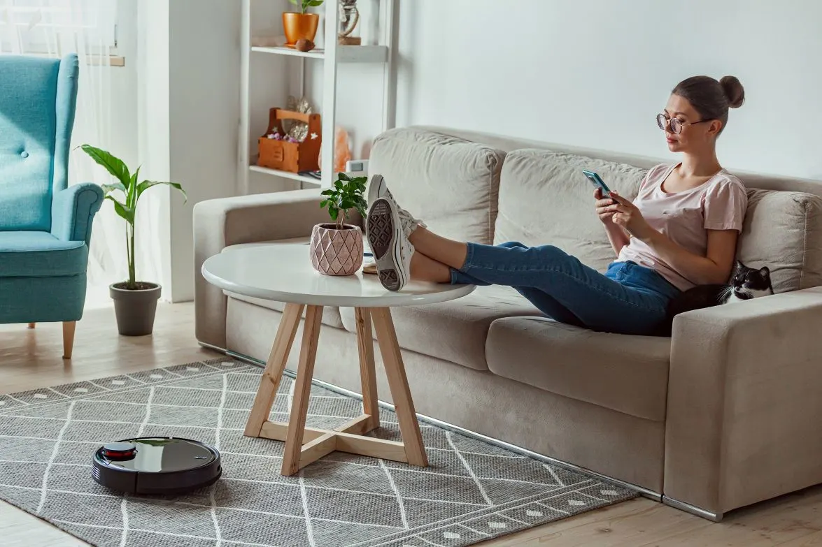 Eine junge Frau mit Brille und Dutt sitzt entspannt auf einem hellen Sofa, hat die Beine auf einem runden weißen Couchtisch abgelegt und bedient ein Smartphone. Neben ihr auf dem Sofa schaut eine schwarz-weiße Katze hervor, während auf dem gemusterten Teppich im Vordergrund ein dunkler Saugroboter fährt.