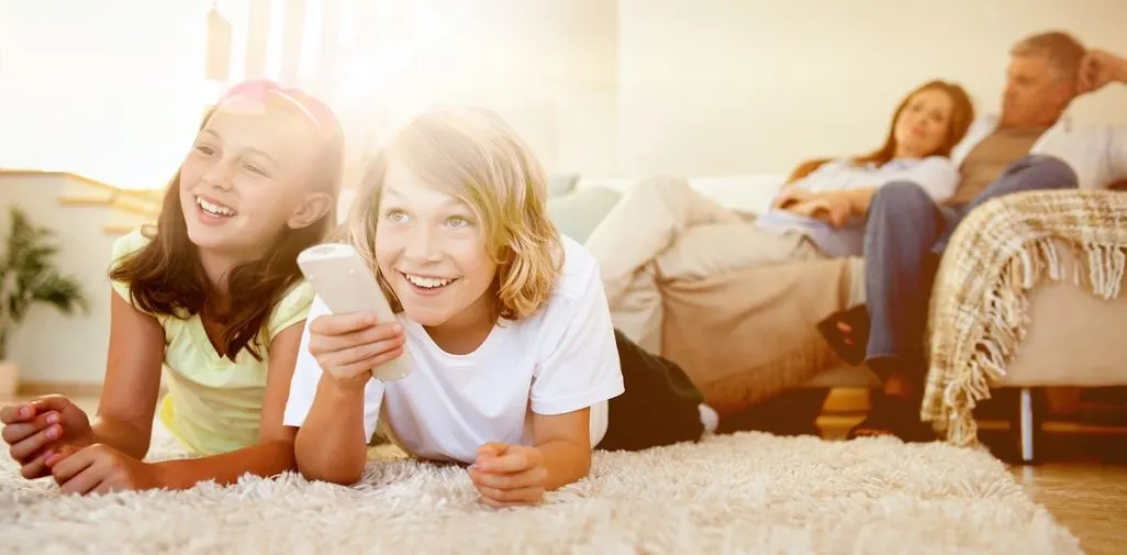 Zwei Kinder liegen auf dem Teppich mit Fernbedienung, Erwachsene sitzen im Hintergrund auf Sofa