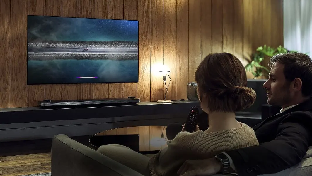 Zwei Personen auf einem grauen Sofa sehen einen Fernseher mit einer dunklen Landschaft, Straße und Reflexionen. Eine Soundbar, Lampe und Holzwand sind sichtbar.