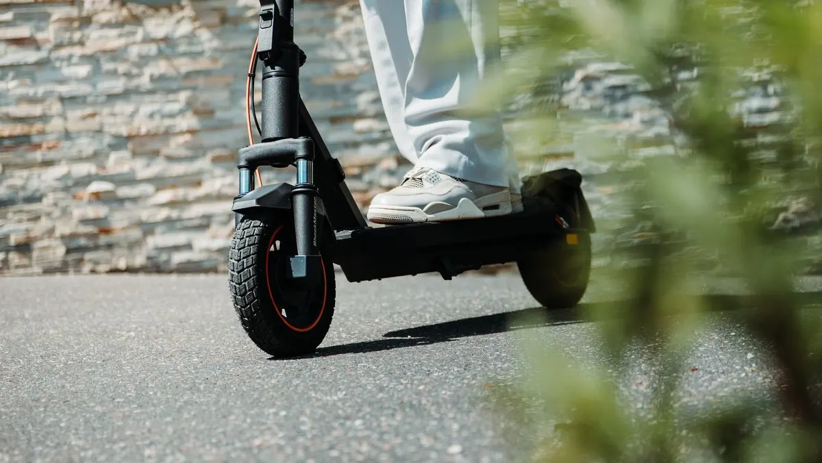 Eine Person steht mit Sportschuhen auf einem E-Scooter und fährt auf einer Straße.