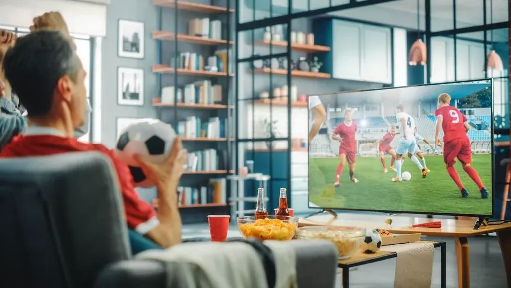In einem Wohnzimmer sitzen zwei Personen auf der Couch und fiebern bei einem Fußballspiel im TV mit. Am Tisch stehen eine Schüssel Chips und Getränkeflaschen.