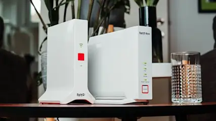 Zwei weiße Fritz!Box Geräte mit grünen Lichtern und roten Knöpfen auf dunklem Holztisch. Ein strukturiertes Wasserglas steht daneben. Unscharfe Pflanzen im Hintergrund.