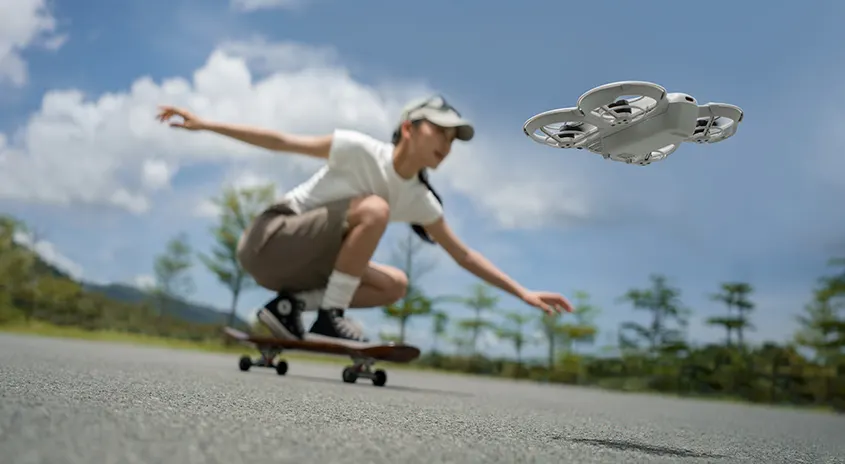 Ein weißer Quadcopter schwebt über einer grauen Asphaltstraße. Ein Skateboard befindet sich auf der Straße vor Bäumen und blauem Himmel.