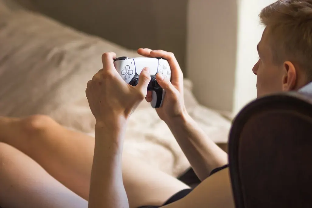 Person sitzt auf dem Bett und hält einen weißen PlayStation Controller in beiden Händen