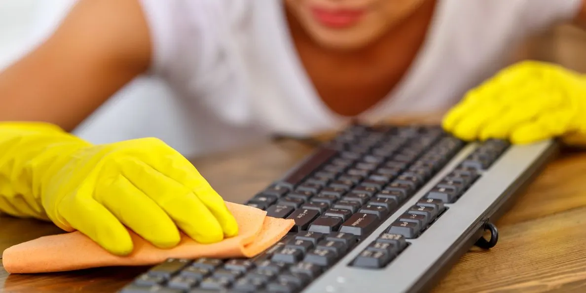 Eine Person mit gelben Handschuhen reinigt eine schwarze Tastatur mit einem orangefarbenen Tuch auf einem Holzschreibtisch.