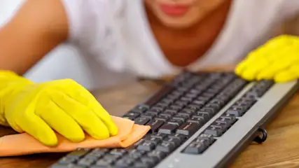 Eine Person mit gelben Handschuhen reinigt eine schwarze Tastatur mit einem orangefarbenen Tuch auf einem Holzschreibtisch.