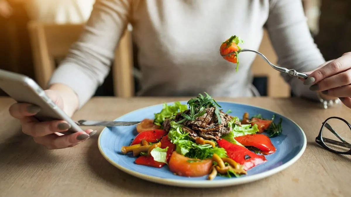 Person sitzt am Tisch, hält in der einen Hand ein Smartphone und isst mit der anderen Hand Salat mit Tomaten und Fleisch.