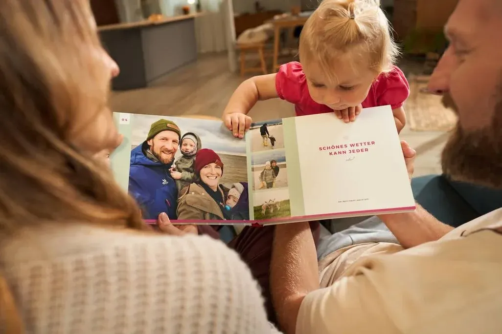 Zwei Erwachsene sitzen nebeneinander und halten ein aufgeschlagenes Fotobuch von CEWE; ein Kleinkind beugt sich vor und schaut auf die Seiten.